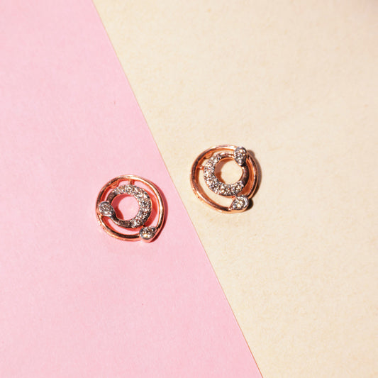 "Rose gold circular earrings with diamond accents arranged on a diagonally split pink and beige background, from jnjbymadhuri."
