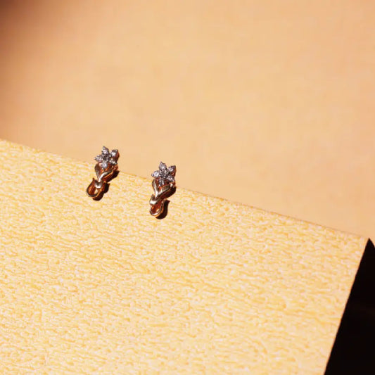 A pair of elegant diamond-studded earrings from jnjbymadhuri, resting on a textured beige surface, illuminated by warm natural light. The delicate sparkle and refined craftsmanship highlight the brand’s dedication to timeless elegance and subtle luxury.
