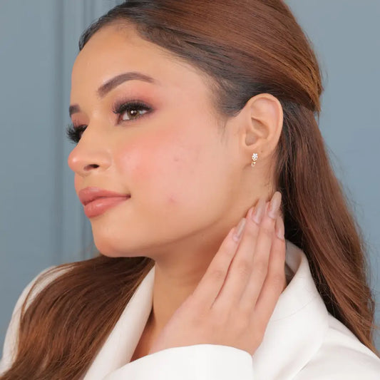 A woman with softly styled, straight brown hair is seen in a side profile, wearing a white blazer and displaying a minimal stud earring. Her hand gently touches her neck, drawing focus to the earring. She wears natural glam makeup with rosy blush and mauve-toned eyeshadow. The image captures a refined and elegant look, perfectly aligned with the jnjbymadhuri jewelry brand's timeless sophistication.