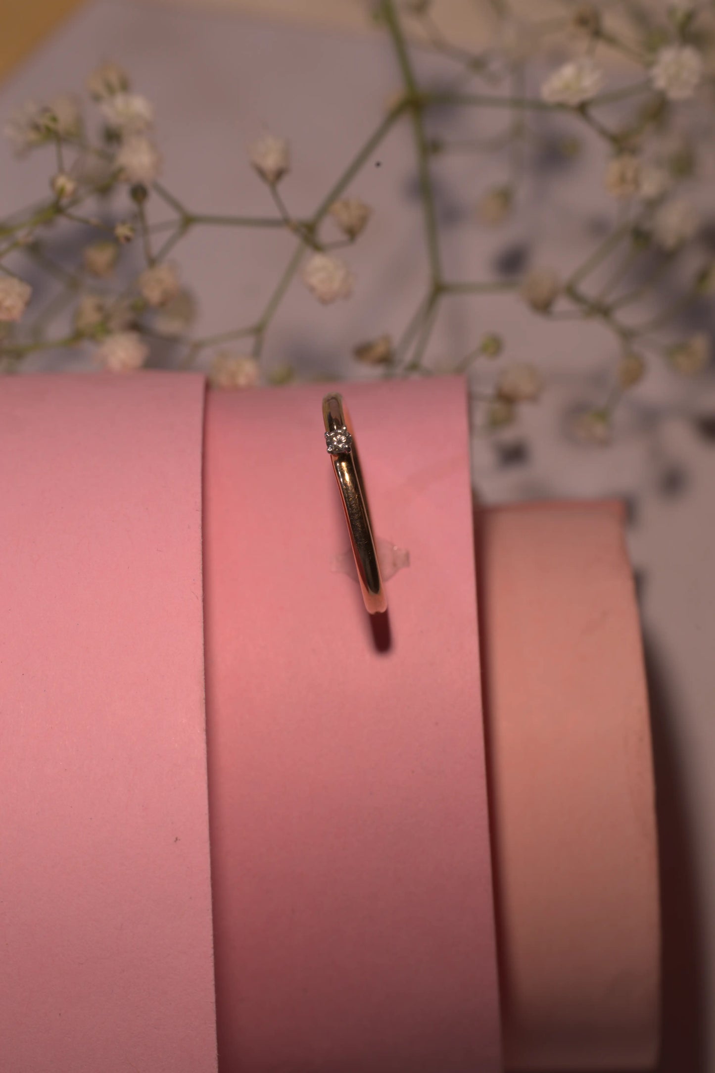 "Delicate jnjbymadhuri ring displayed on a pink cylindrical stand with soft baby’s breath flowers in the background."
