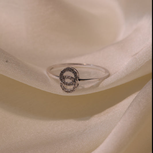 An elegant silver ring with a shimmering circular motif encrusted with delicate stones, showcased on a soft ivory satin fabric, highlighting its refined design and sophisticated appeal by jnjbymadhuri.