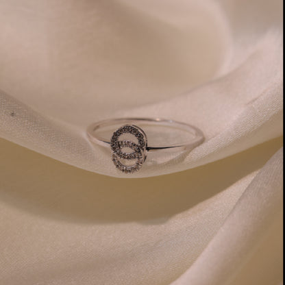An elegant silver ring with a shimmering circular motif encrusted with delicate stones, showcased on a soft ivory satin fabric, highlighting its refined design and sophisticated appeal by jnjbymadhuri.
