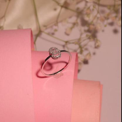 A delicate silver ring with a round, flower-inspired diamond cluster centerpiece is elegantly displayed on soft pink rolled paper, exuding feminine charm and timeless elegance by jnjbymadhuri.
