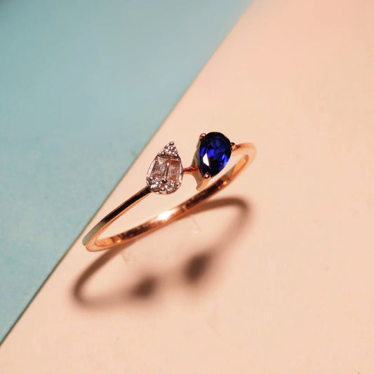 Stylish rose gold open ring from jnjbymadhuri featuring a teardrop-shaped sapphire and a sparkling diamond, artistically displayed on a pastel background.
