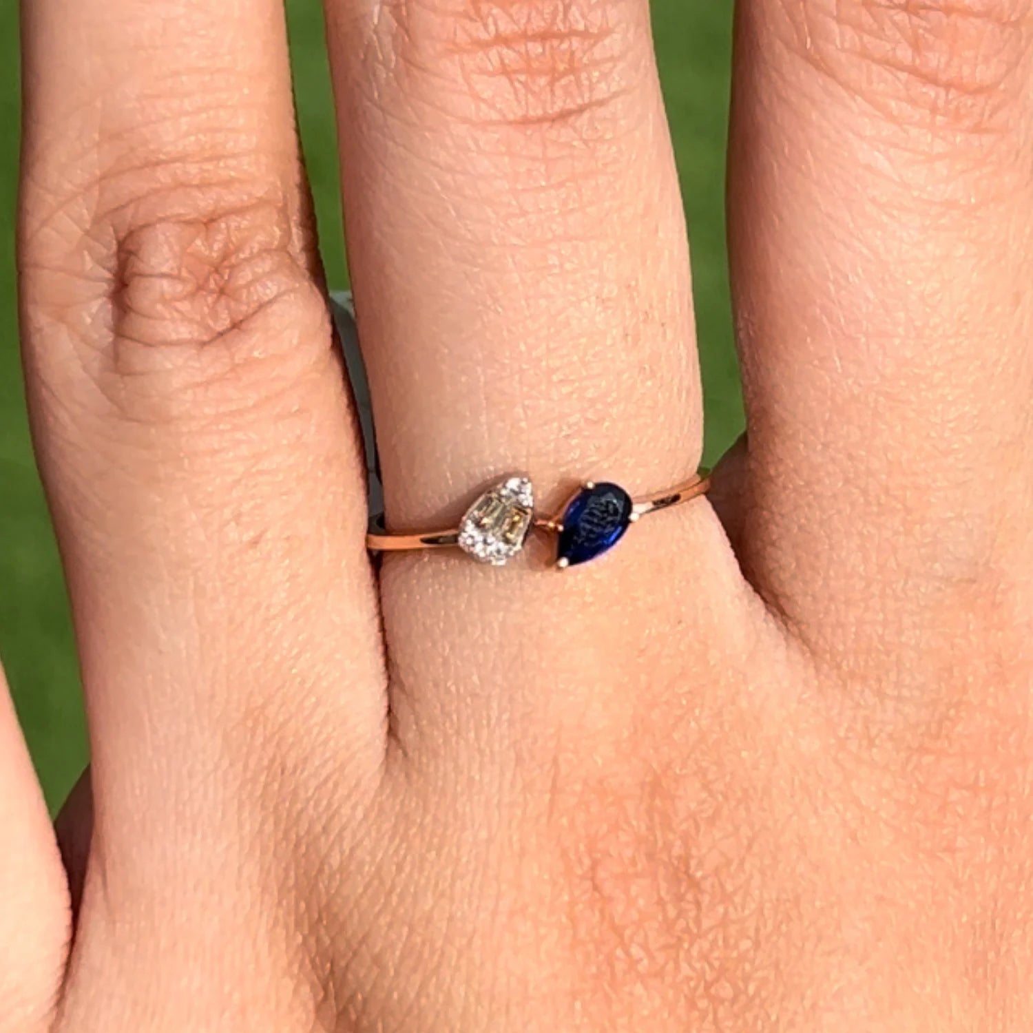 Elegant rose gold ring from jnjbymadhuri featuring a sapphire and diamond teardrop design, gracefully worn on a finger with a natural green background.
